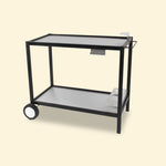 Chariot de plancha en métal noir avec plateaux en inox, doté de deux roues arrière, d’un porte-ustensiles latéral et d’un support à bouteilles intégré, présenté sur fond uni clair.