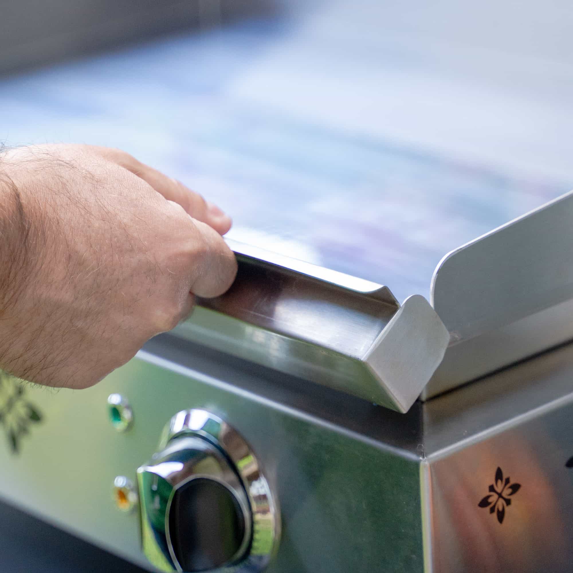 Gros plan sur la main d’une personne manipulant le rebord de la plaque en inox d’une plancha électrique Vivaplancha, avec le bouton de réglage visible au premier plan.