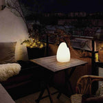 Lampe solaire d’extérieur en forme d’œuf, allumée sur une table en bois sur une terrasse de nuit, diffusant une lumière chaude dans une ambiance cosy.
