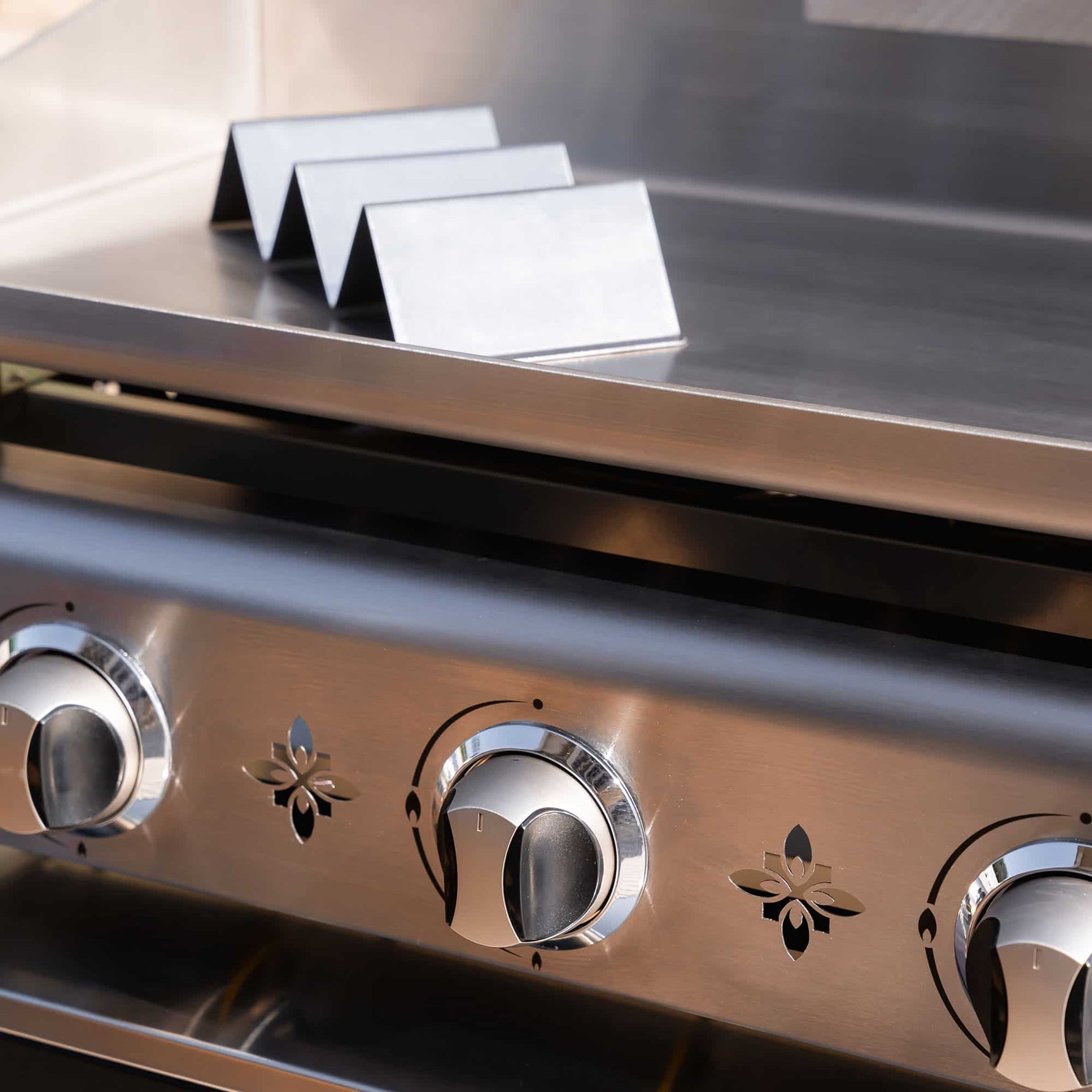 Supports de maintien au chaud en inox posés sur la plaque d’une plancha inox à trois brûleurs, présentés en gros plan en extérieur.